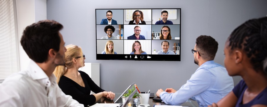 Group of four employees sitting at a conference table, looking at online meeting attendees on the display screen.