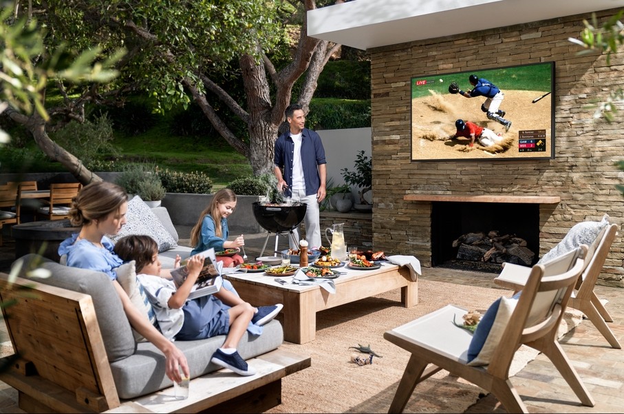 A family watching a baseball game on an outdoor TV on their patio while dad cooks food on a small grill. 