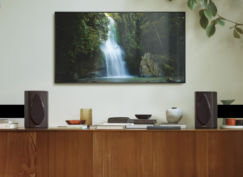 A wall-mounted TV displaying a waterfall scene, flanked by a pair of Sonus Faber Duetto speakers, on a wooden counter. 